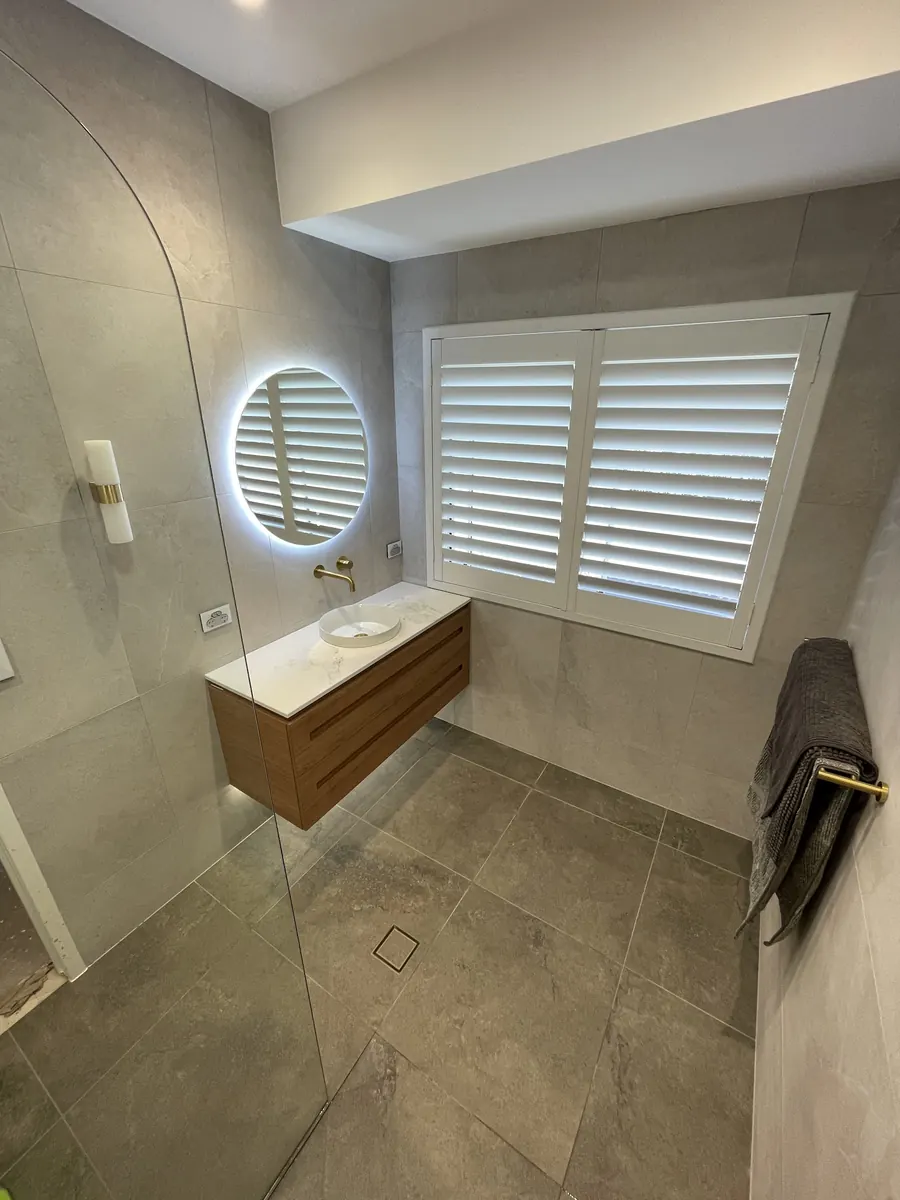 Bathroom with a round backlit mirror, timber vanity, plantation shutters and a frameless shower