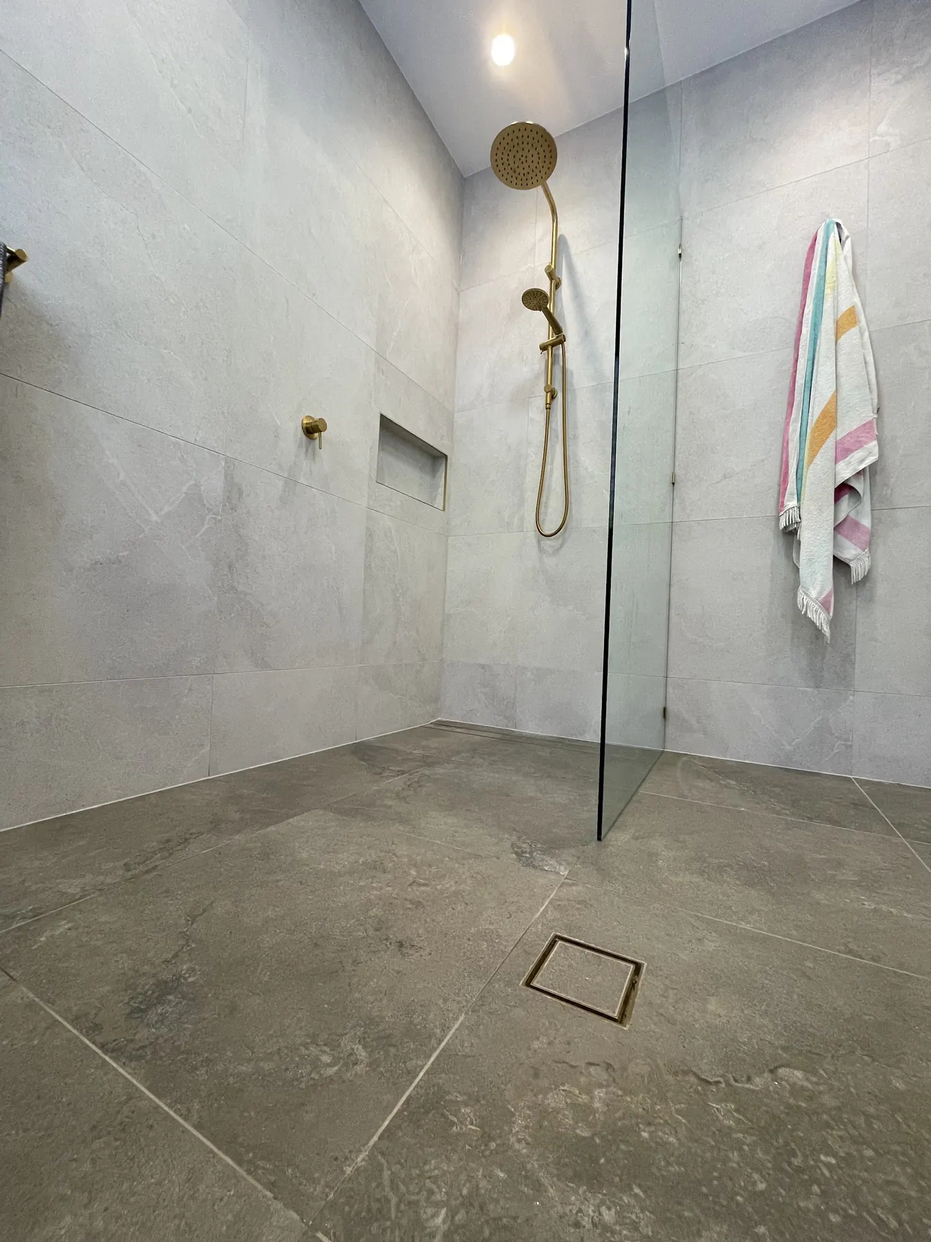 Walk-in shower tiled in large-format grey tiles with a brass floor drain and brass shower rail